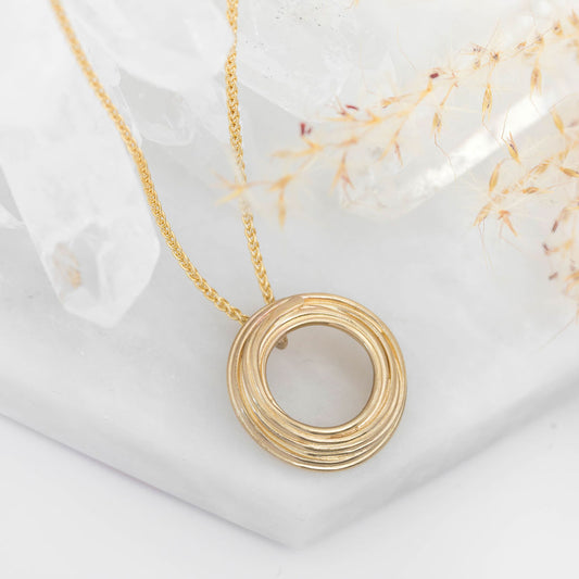 Gold necklace with a circular pendant on a white surface with dried flowers. Handcrafted by Angela Evans.